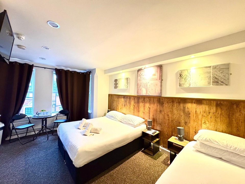 Spacious guest room with a double and a single bed along an oak-panelled wall, a table with two chairs by the windows and a wall-mounted TV